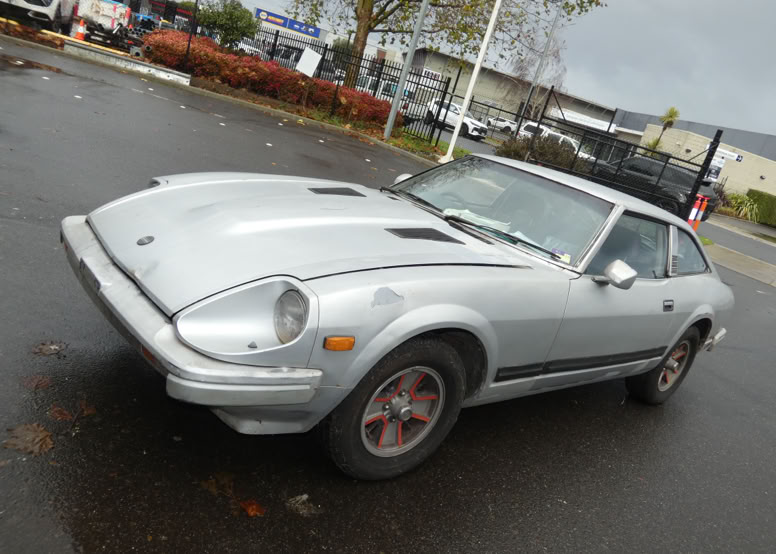 A silver vintage sports car parked in a parking lot on a cloudy day.