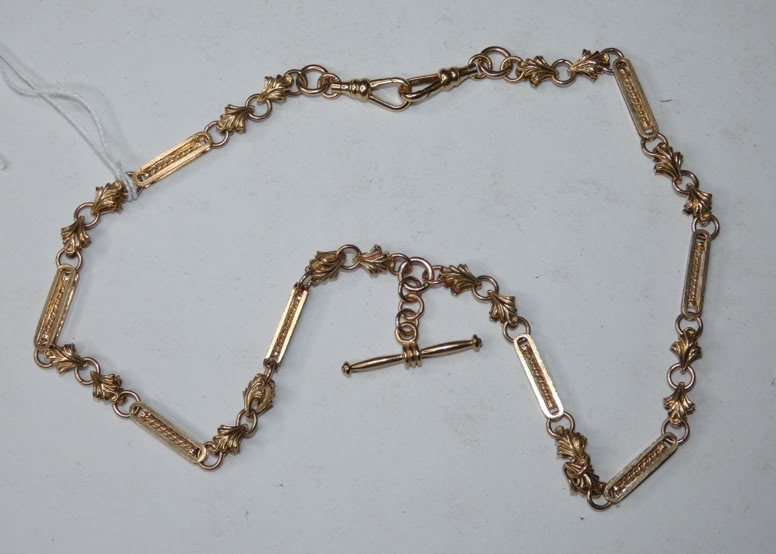 A gold chain with rectangular and circular links, featuring a T-bar clasp, laid out on a white surface.