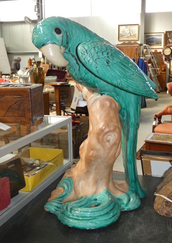 A green ceramic parrot perched on a decorative stand in an antique shop setting.